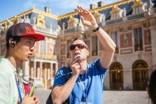 Tour guide gestures outside a historic building, speaking to a tourist wearing headphones.