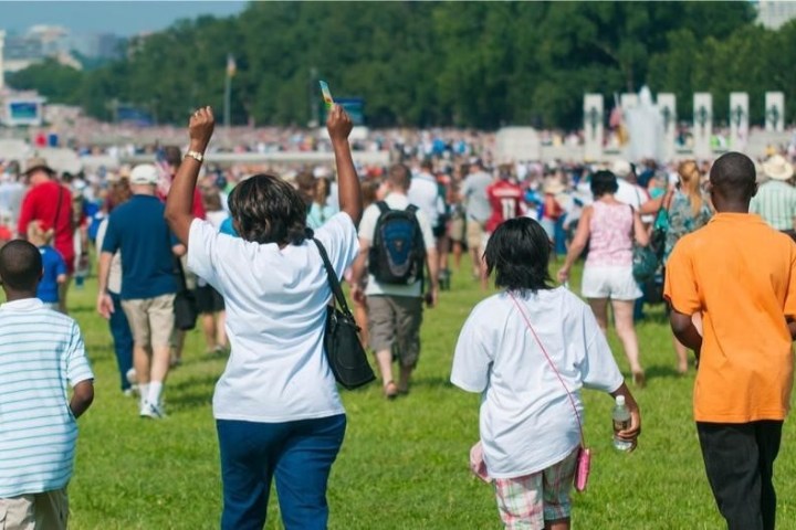 Crowd walking in a park towards a monument on a sunny day.
