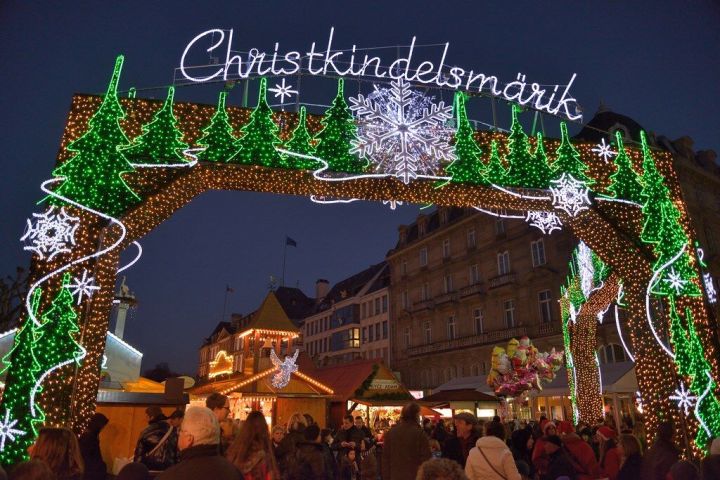 Festive Christmas market arch with lights, trees, and snowflakes at night, crowded with people.