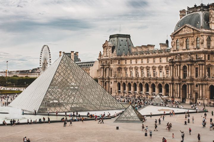 Glass pyramid at the Louvre Museum courtyard, with Ferris wheel and historic building in the background.