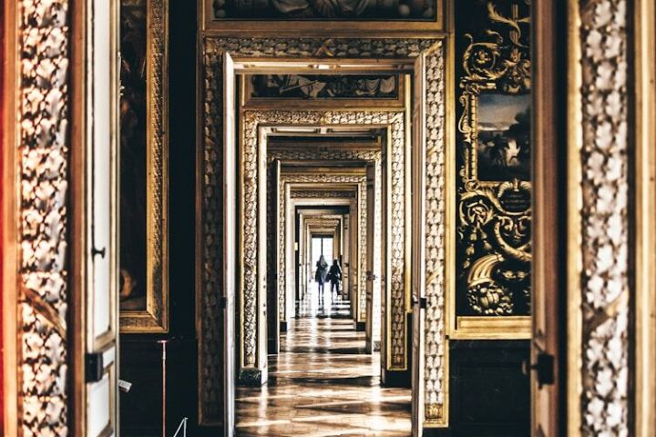 Ornate hallway with repeating door frames, sunlight on parquet floor, and distant figures.