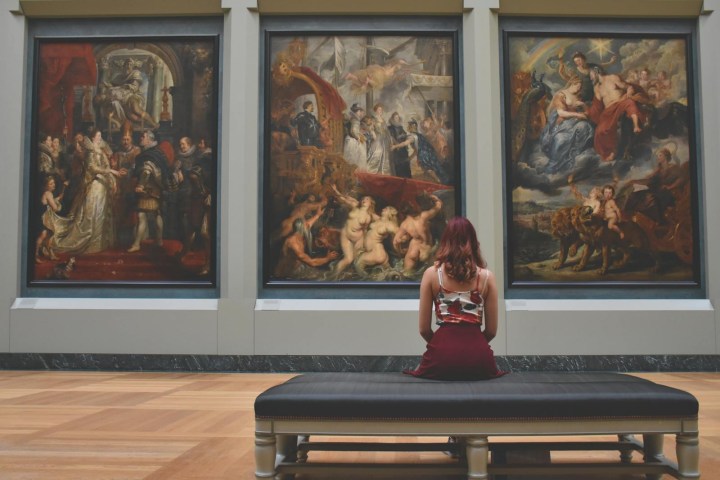 Woman sitting on bench, viewing three large paintings in a museum.