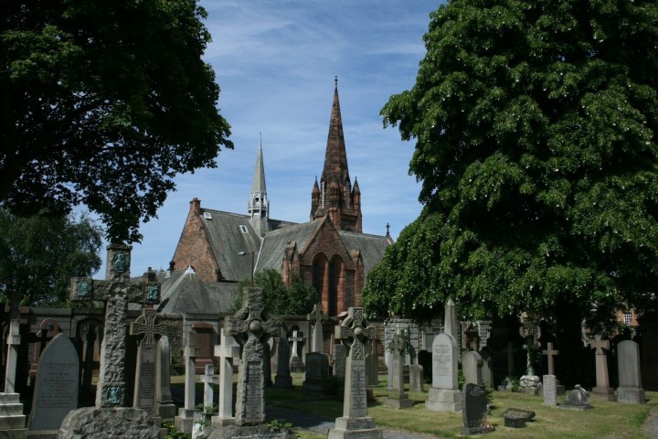 Cemetery with tombstones and trees, old church with spires in the background against blue sky.
