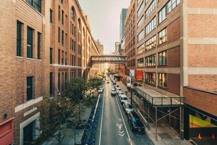Urban street with brick buildings, parked cars, bicycles, and a sky bridge between buildings.