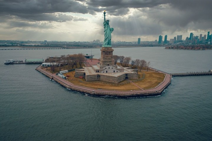 Aerial view of the Statue of Liberty on Liberty Island with NYC skyline, cloudy sky.
