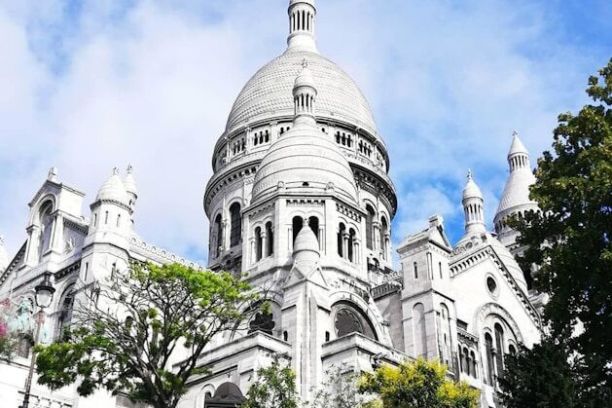 White domed basilica with stairs, lush greenery, and blue sky with clouds.