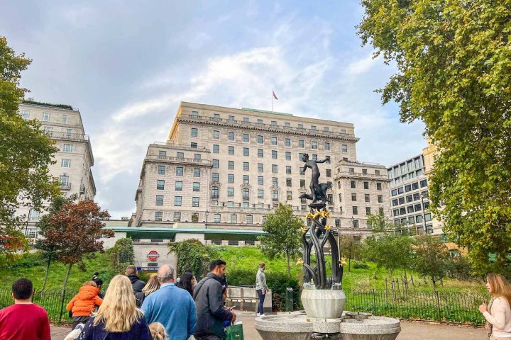 People walking near fountain with tall city building in background on a cloudy day.