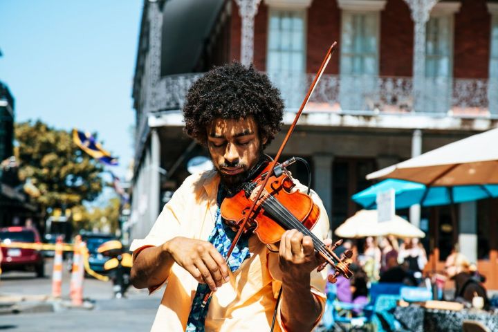Man playing violin outdoors on a sunny street with people in the background.