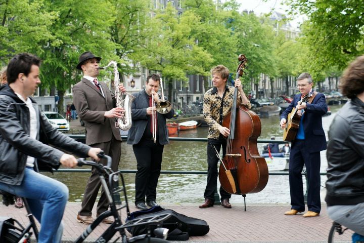 Street musicians play saxophone, trombone, double bass, and guitar near a canal with cyclists passing by.