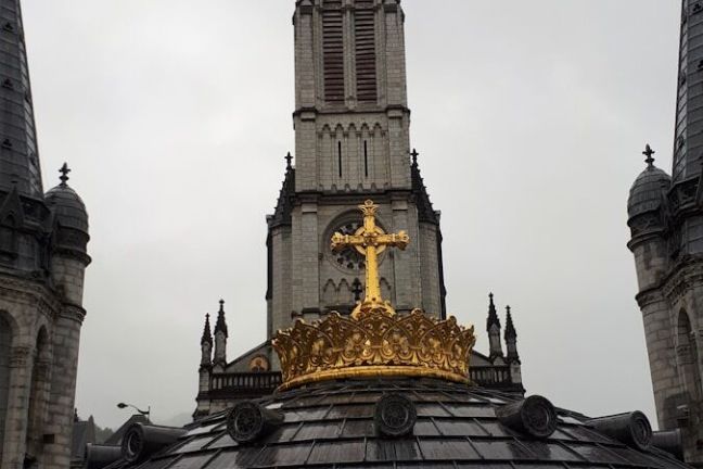 Cathedral tower with gold cross on domed roof, overcast sky.