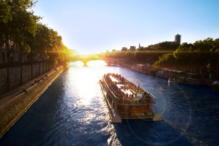 Tour boat on a river in the evening sun, with trees and bridge in the background.