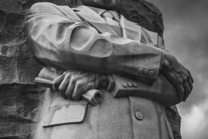 Stone statue of a man with arms crossed, wearing a suit, against a cloudy sky backdrop.