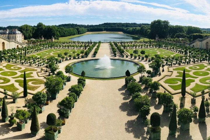 Panoramic view of symmetrical gardens with fountain and ornate patterns, surrounded by trees and a long canal.