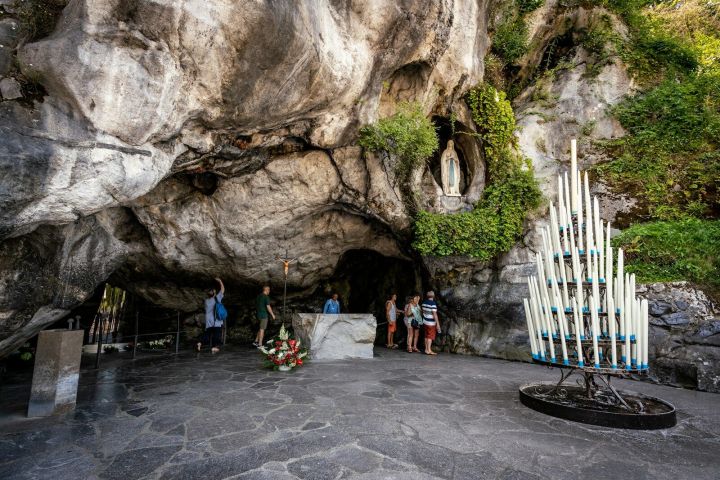 People at a grotto with a statue in the niche and a stand of large candles in front.