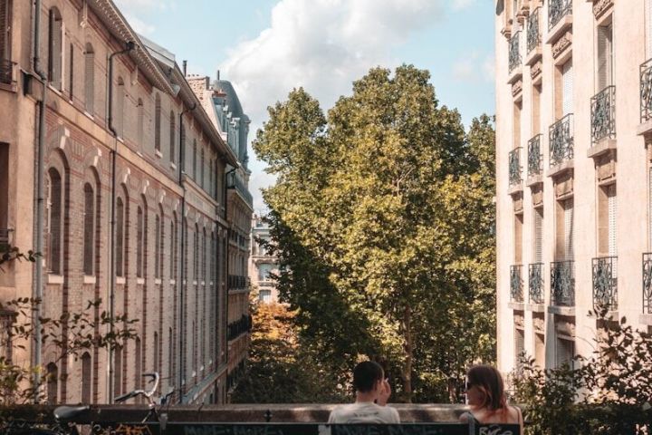 Two people sitting on a bench in a narrow street with trees and buildings, under a blue sky with clouds.