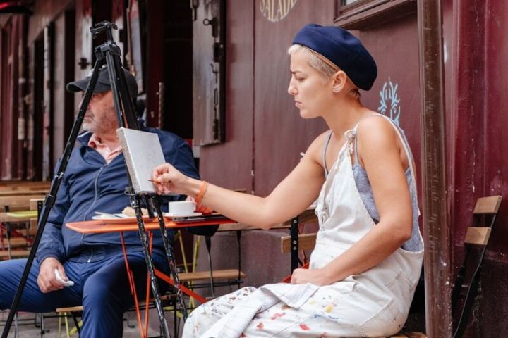 Woman in apron and beret painting outdoors at a cafe table, with a man sitting nearby.