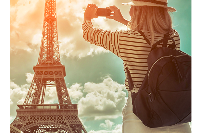 Woman photographing Eiffel Tower with a smartphone, wearing a hat and backpack.