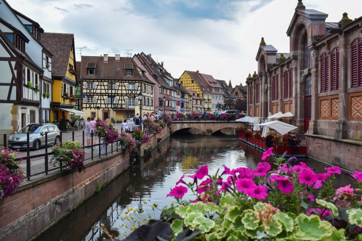 Colorful historic European street with canal, flowers, and bridge under a cloudy sky.