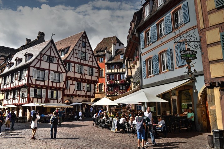 Charming European street with half-timbered buildings, outdoor cafes, and people walking in sunlight.