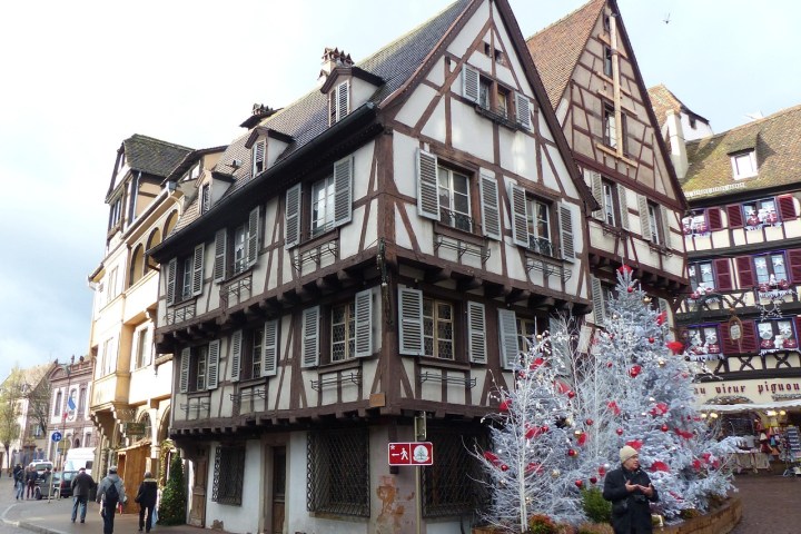 Historic half-timbered houses in a European street, with decorated Christmas trees and people walking nearby.