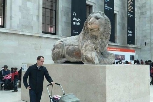 Man with stroller walks by a large lion statue in a museum atrium.