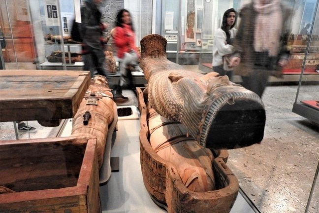 Museum exhibit of ancient Egyptian sarcophagi with blurred visitors walking by.