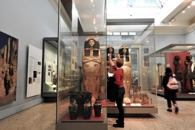 Museum exhibit with sarcophagi in glass cases and visitors observing.