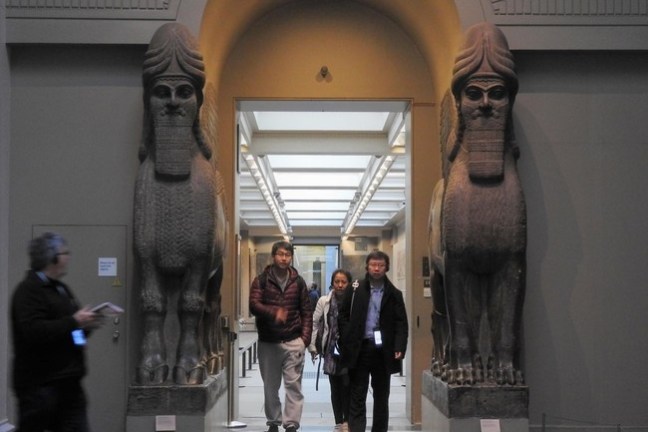 Three people walk between large ancient statues in a museum.