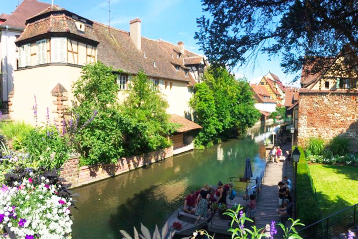 Scenic canal with traditional houses, lush greenery, and people enjoying a boat ride on a sunny day.