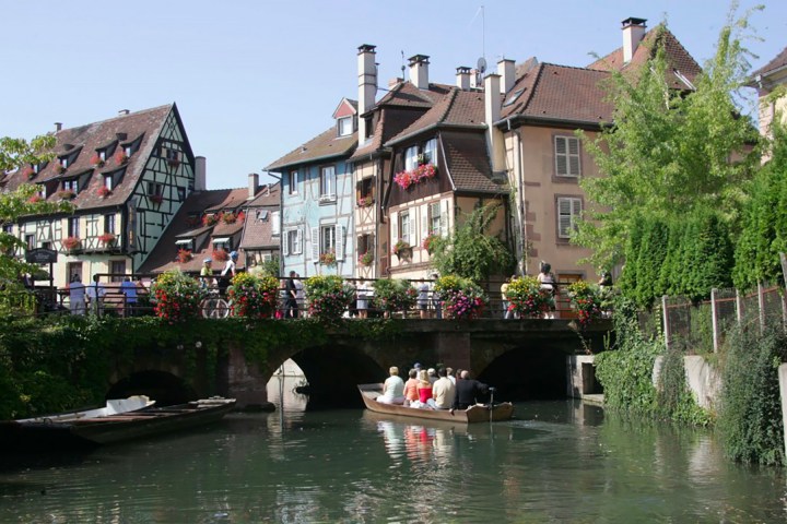 Charming canal scene with people in a boat, surrounded by colorful, half-timbered buildings and lush greenery.