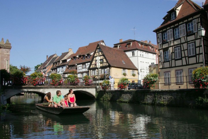 People on a boat in a canal with half-timbered houses and a bridge in the background.