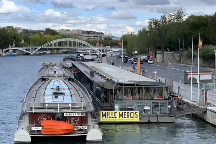 Boat docked on river with pedestrian bridge and trees in background under a cloudy sky.