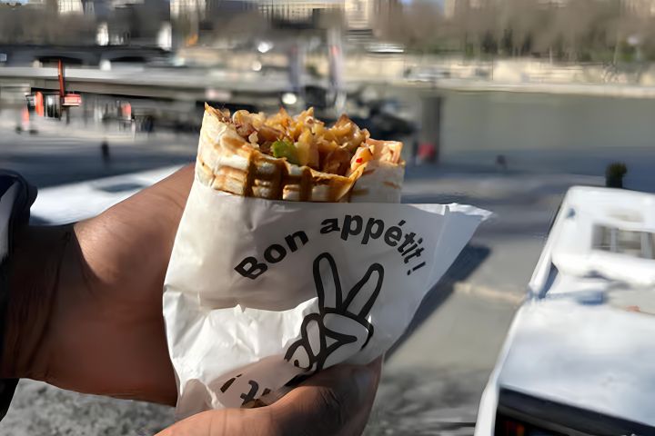 Hand holding a wrapped sandwich with 'Bon appétit!' near a river.