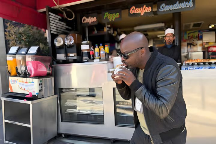 Man eating a sandwich in front of a street food stand with drinks and snacks.