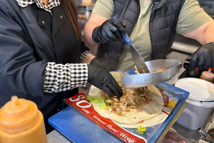 Two people assembling a wrap with tongs and a spatula over a blue cutting board.
