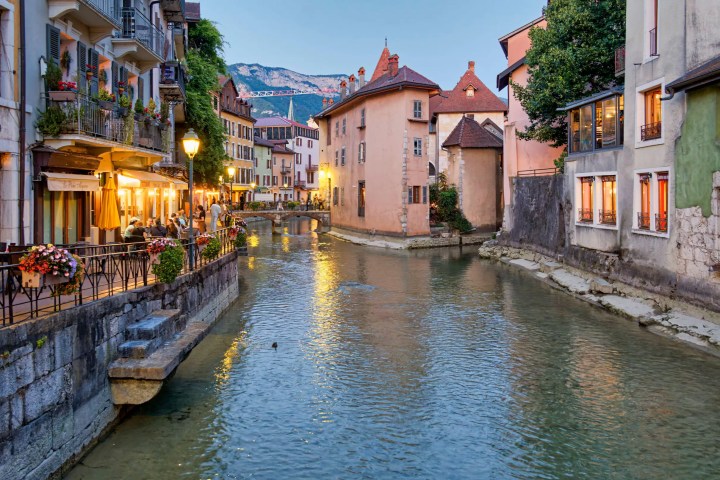 Charming canal with historic buildings and outdoor cafe at dusk, surrounded by lush greenery.