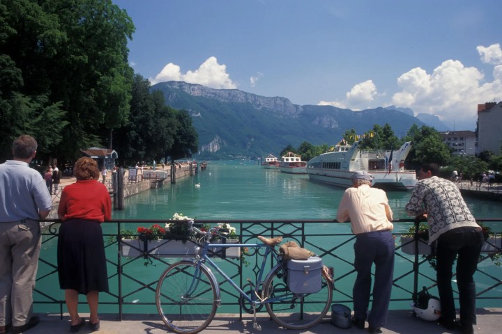 People on a riverside bridge with mountains and boats in the background.
