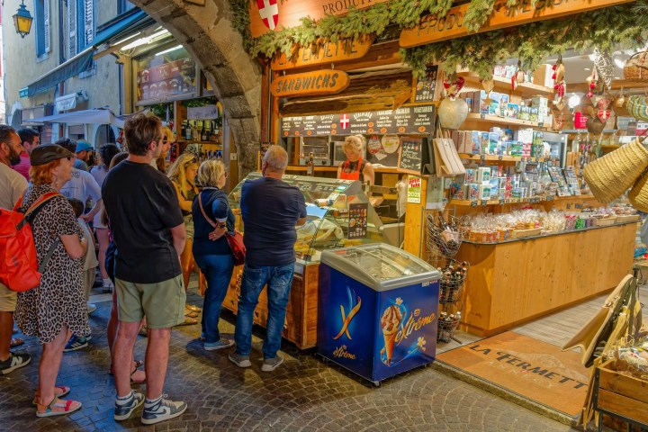 People lined up at a small shop with wooden facade under an archway.