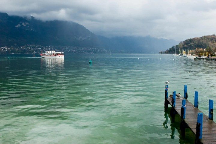 Lake with boat, pier, and mountains under cloudy sky.