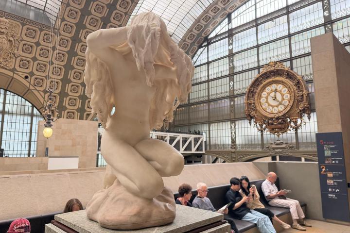 Kneeling sculpture with drapery in art museum, large clock in background.