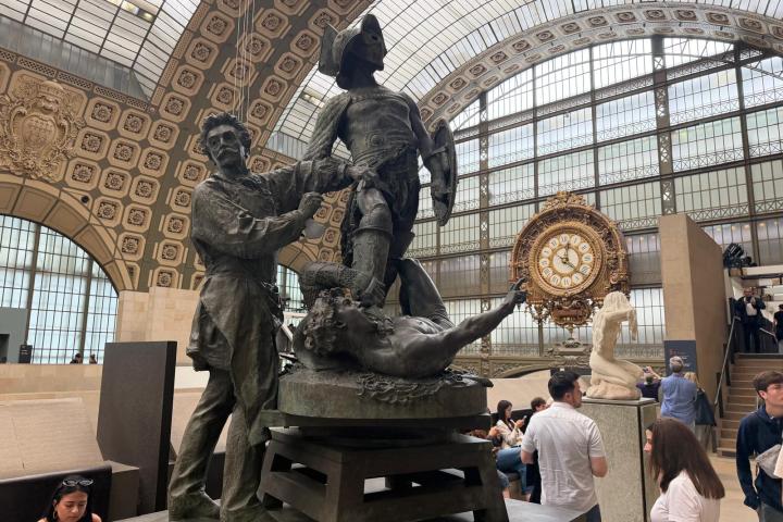 Statue inside a grand hall with a large clock and people around.