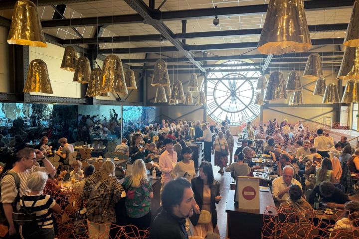 Crowded café with large clock window and gold lamps, people dining and mingling.